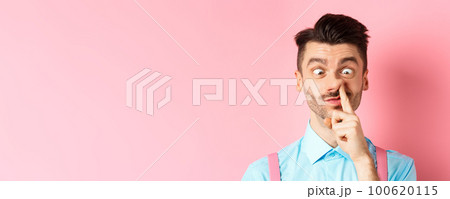 Image of funny caucasian guy picking nose and squint eyes, making dumb face, standing on pink background 100620115