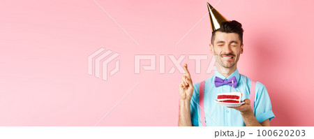 Holidays and celebration concept. Hopeful birthday guy in party hat making wish, holding cake with candle and cross fingers for dream come true, pink background 100620203