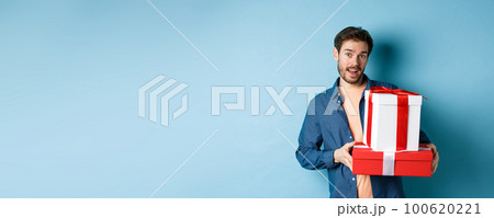 Handsome bearded guy holding Valentines day gifts for lover, standing with presents in boxes and looking at camera, blue background 100620221