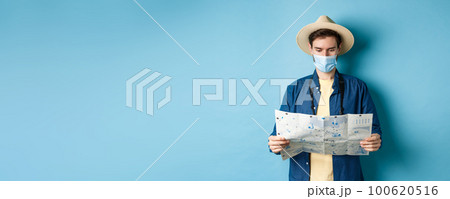 Covid-19, pandemic and travel concept. Young man in medical mask reading tourist map, searching for route to sightseeings, standing on blue background 100620516