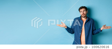 Indecisive young man with beard, look away and shrugging shoulders, know nothing, being clueless, standing on blue background Indecisive young man with beard, look away and shrugging shoulders, know nothing, being clueless, standing on blue background 100620616