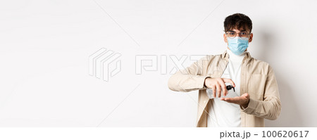 Health, covid and quarantine concept. Young hispanic guy in glasses and face mask using hand sanitizer, apply antiseptic and look at camera, standing on white background Health, covid and quarantine concept. Young hispanic guy in glasses and face mask using hand sanitizer, apply antiseptic and look at camera, standing on white background 100620617