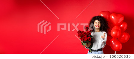Holidays and celebration. Beautiful girlfriend receive flowers on anniversary, showing finger heart and standing near party balloons, red background Holidays and celebration. Beautiful girlfriend receive flowers on anniversary, showing finger heart and standing near party balloons, red background 100620639