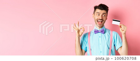 Cheerful man recommending great promo, showing OK sign and looking excited at plastic credit card, standing on pink background 100620708