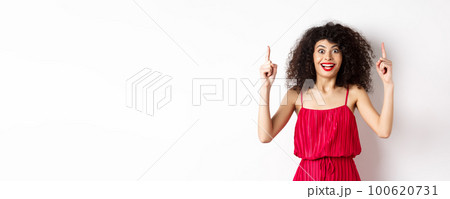 Happy elegant woman in red dress and makeup, smiling amused and pointing fingers up at logo, showing advertisement, standing over white background 100620731