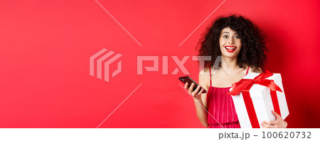 Online shopping and Valentines day. Beautiful young woman holding smartphone and lovers gift, looking surprised and happy at camera, red background 100620732