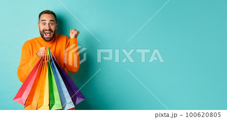 Lifestyle concept. Excited man showing shopping bags and rejoicing from discounts, standing over turquoise background 100620805