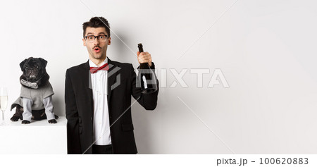 Pets, winter holidays and New Year concept. Cheerful man with cute black pug dog celebrating Christmas party, holding champagne bottle and smiling, white background Pets, winter holidays and New Year concept. Cheerful man with cute black pug dog celebrating Christmas party, holding champagne bottle and smiling, white background 100620883