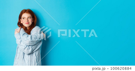 Happy cute redhead girl hugging herself, wearing comfortable and warm sweater, smiling at camera, standing over blue background Happy cute redhead girl hugging herself, wearing comfortable and warm sweater, smiling at camera, standing over blue background 100620884