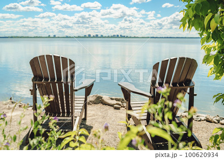 Two Muskoka chairs by the water on home terrace with calm view of lake in Canada. Summer cottage vacation lifestyle 100620934