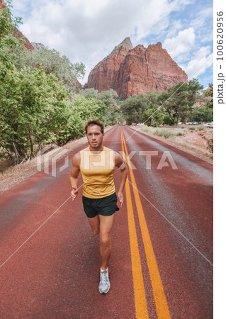Athlete runner running on outdoor run jogging on red road outside in nature landscape. Vertical portrait of fit healthy young man training cardio for marathon or triathlon. 100620956