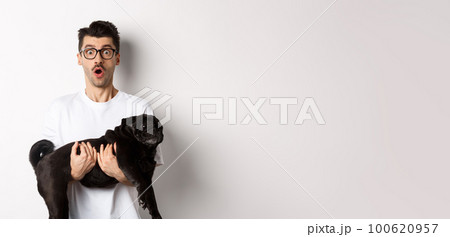 Surprised young man in glasses holding cute black pug, dog owner staring at camera with impressed face, saying wow, standing over white background Surprised young man in glasses holding cute black pug, dog owner staring at camera with impressed face, saying wow, standing over white background 100620957