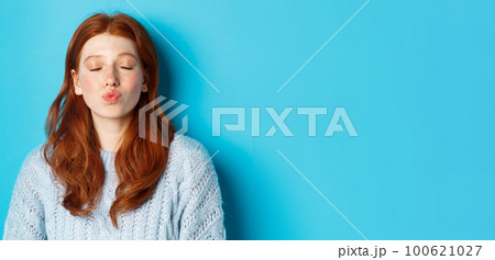 Close-up of young woman with red wavy hair standing with eyes closed and puckered lips, waiting for kiss against blue background 100621027