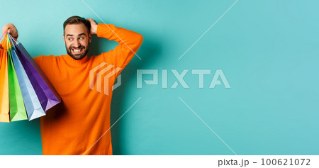 Handsome young man looking awkward and scratching head, staring at shopping bags with gifts, standing over turquoise background 100621072