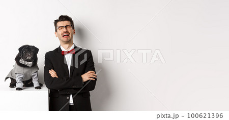 Animals, party and celebration concept. Sad dog owner crying, wearing suit, standing near cute black pug in costume, standing over white background 100621396