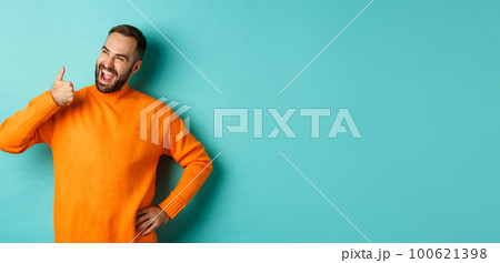 Happy satisfied male model showing thumb up, looking pleased at upper left corner logo, approve and recommend product, standing over light blue background 100621398