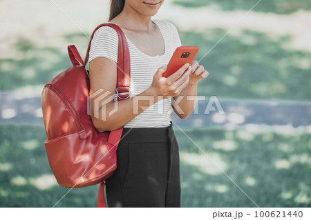 Woman using phone outside walking in park carrying red bag on autumn back to school campus. Online app for vaccine passport. Woman using phone outside walking in park carrying red bag on autumn back to school campus. Online app for vaccine passport. 100621440