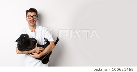 Handsome young man playing with cute black puppy. Dog owner petting a pug, standing over white background 100621469