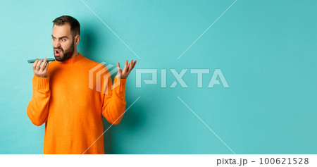 Angry man arguing on speakerphone, record voice message with mad face, standing over light blue background in orange sweater 100621528
