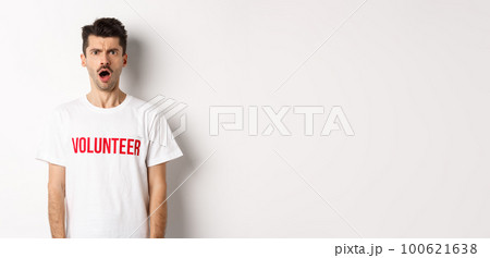 Shocked and confused man in volunteer t-shirt staring at camera and frowning displeased, standing against white background 100621638