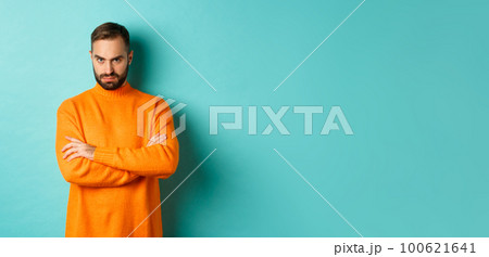 Offended man looking angry at you, cross arms on chest and stare mad, standing in orange sweater against turquoise background 100621641