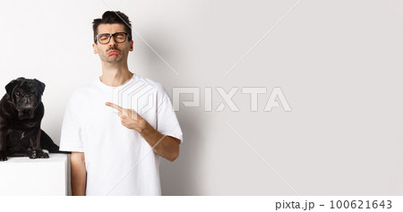 Sad and gloomy pet owner pointing at his black pug dog and sobbing, standing upster against white background 100621643