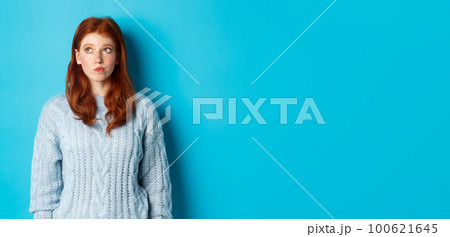 Dreamy redhead girl thinking or making decision, looking at upper left corner logo, standing against blue background 100621645