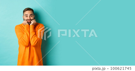 Sad man sighing and looking thoughtful, standing in orange sweater against turquoise background 100621745