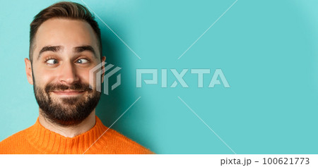 Headshot of funny caucasian man making faces, squinting and smiling silly, standing over light blue background 100621773