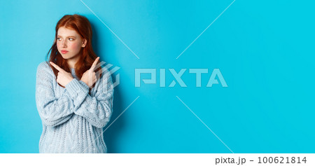 Indecisive redhead teenage girl making decision, pointing fingers sideways and looking left doubtful, standing in sweater against blue background 100621814