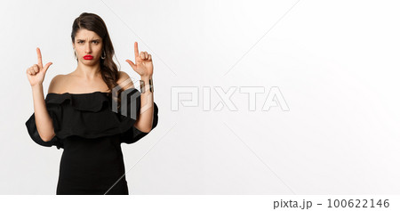 Fashion and beauty. Disappointed woman sulking upset, pointing fingers up and complaining, standing dissatisfied in black dress, white background Fashion and beauty. Disappointed woman sulking upset, pointing fingers up and complaining, standing dissatisfied in black dress, white background 100622146