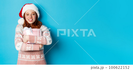 Winter and celebration concept. Dreamy redhead girl in santa hat hugging her christmas gift, smiling happy, standing over blue background Winter and celebration concept. Dreamy redhead girl in santa hat hugging her christmas gift, smiling happy, standing over blue background 100622152