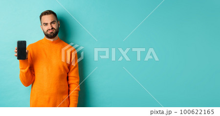 Displeased and disappointed man in orange sweater, grimacing and showing mobile screen, standing over light blue background 100622165