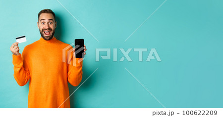 Online shopping. Excited man showing mobile screen and credit card, smiling amazed, standing over turquoise background 100622209