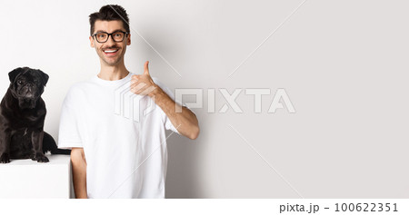 Image of dog owner and cute black pug looking at camera, man showing thumb-up in approval, recommending something, standing over white background Image of dog owner and cute black pug looking at camera, man showing thumb-up in approval, recommending something, standing over white background 100622351