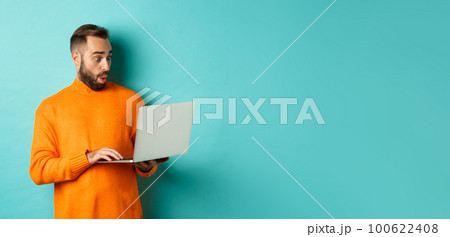 Freelance and technology concept. Image of handsome adult man staring at laptop with amazement, shopping online or working remote, standing over turquoise background Freelance and technology concept. Image of handsome adult man staring at laptop with amazement, shopping online or working remote, standing over turquoise background 100622408