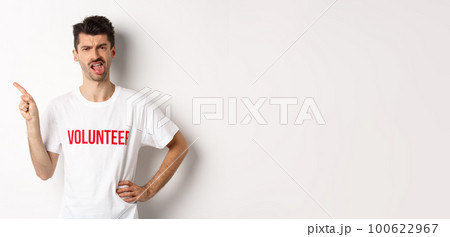 Disappointed volunteer showing bad promo offer, sticking tongue and grimacing disgusted, pointing finger left, wearing organization t-shirt, white background Disappointed volunteer showing bad promo offer, sticking tongue and grimacing disgusted, pointing finger left, wearing organization t-shirt, white background 100622967