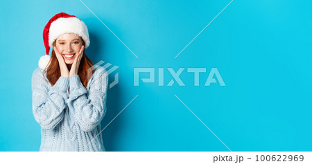 Winter holidays and Christmas Eve concept. Cheerful redhead girl in santa hat, celebrating New Year, blushing and smiling happy, standing over blue background 100622969