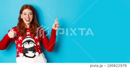 Merry Christmas. Cheerful redhead girl in xmas sweater, pointing finger at upper right corner, showing new year promo and thumbs-up in approval, praise product Merry Christmas. Cheerful redhead girl in xmas sweater, pointing finger at upper right corner, showing new year promo and thumbs-up in approval, praise product 100622976