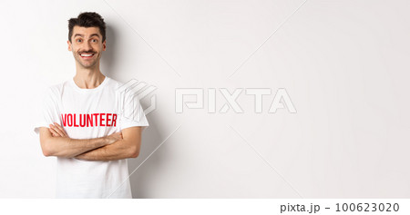 Happy young man in volunteer t-shirt ready to help, smiling at camera, cross arms on chest confident, white background 100623020