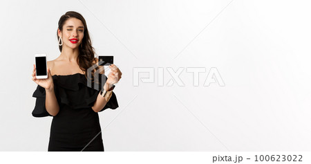Fashion and shopping concept. Beautiful woman with red lips, winking at camera, showing smartphone screen and credit card, buying online, white background Fashion and shopping concept. Beautiful woman with red lips, winking at camera, showing smartphone screen and credit card, buying online, white background 100623022