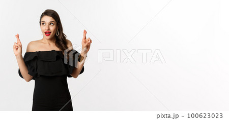 Fashion and beauty. Excited gorgeous woman in black dress, holding fingers crossed and looking at upper left corner, making wish, standing over white background 100623023
