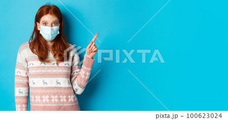 Winter, covid-19 and quarantine concept. Intrigued redhead girl in medical mask picking product, looking and pointing at upper right corner promo, blue background 100623024
