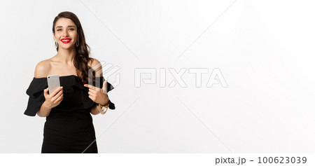 Online shopping concept. Stylish woman in black dress, wearing makeup, pointing finger at mobile phone with satisfied smile, standing over white background Online shopping concept. Stylish woman in black dress, wearing makeup, pointing finger at mobile phone with satisfied smile, standing over white background 100623039