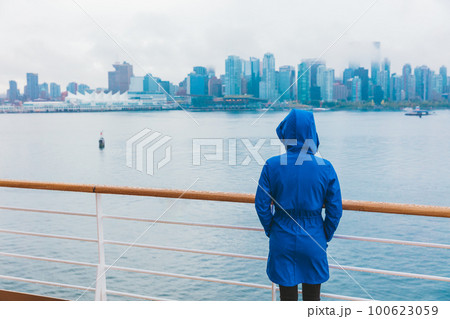 Autumn city scenery woman walking alone under the rain wearing blue raincoat in wet cold day. Skyline of Vancouver, Canada. Cruise ship travel destination. 100623059