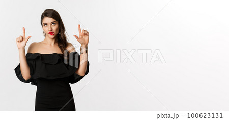 Fashion and beauty. Thoughtful attractive woman in black dress staring and pointing up, thinking with tempted look, white background 100623131