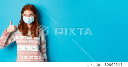Winter, coronavirus and social distancing concept. Pretty redhead girl in face mask and sweater, looking at upper left corner promo and showing thumb up, praising good product Winter, coronavirus and social distancing concept. Pretty redhead girl in face mask and sweater, looking at upper left corner promo and showing thumb up, praising good product 100623134