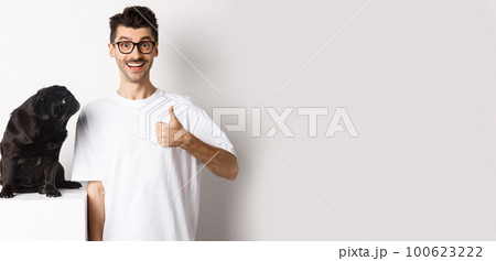 Cheerful handsome guy standing near cute black pug and showing thumb-up. Pet owner approve and recommend product for dogs, white background Cheerful handsome guy standing near cute black pug and showing thumb-up. Pet owner approve and recommend product for dogs, white background 100623222