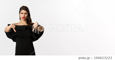 Fashion and beauty. Disappointed and upset woman in black dress, showing thumbs down, dislike something bad, judging over white background 100623223
