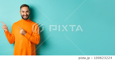 Handsome smiling man pointing fingers left, showing your logo, standing in winter orange sweater, turquoise background Handsome smiling man pointing fingers left, showing your logo, standing in winter orange sweater, turquoise background 100623224